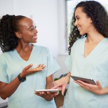 Two people in the medical field wearing scrubs discuss with digital tablets. 