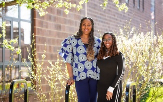 Two people smiling outside of a brick building surrounded by greenery.