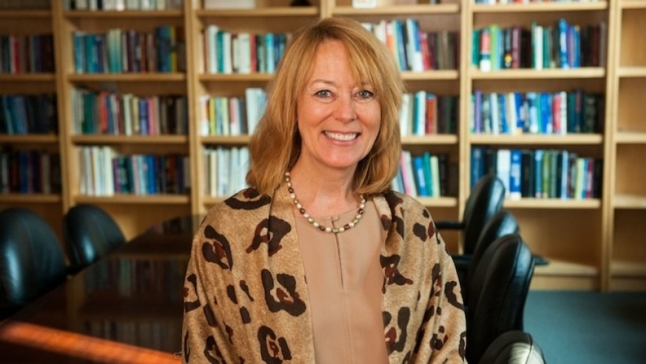 Peggy Compton stands in front of a table and bookcase.
